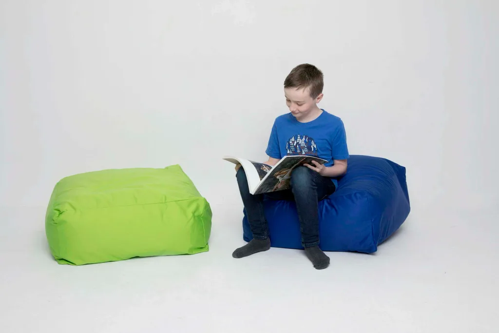 Child sitting on a blue cotton square bean bag reading a book, with a green cotton square bean bag beside.
