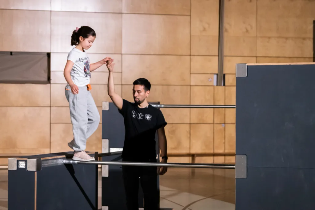 Child balancing on a multiplex birch parkour platform with high-grip surface while an adult instructor holds her hand for support.