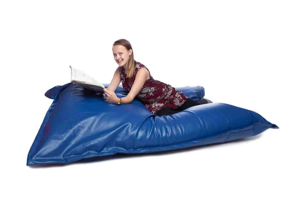 Teenager reclining on a royal blue faux leather floor cushion while reading a book.