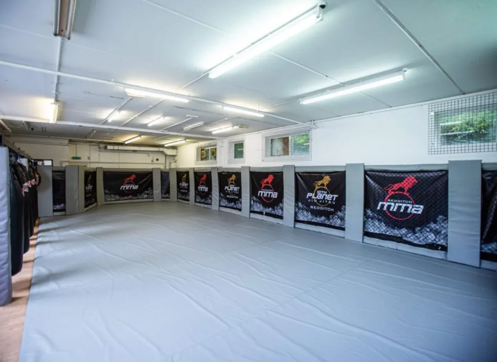 Steel cage walling with powder-coated branded panels reading MMA and grey padded corner posts around a large grey training mat.