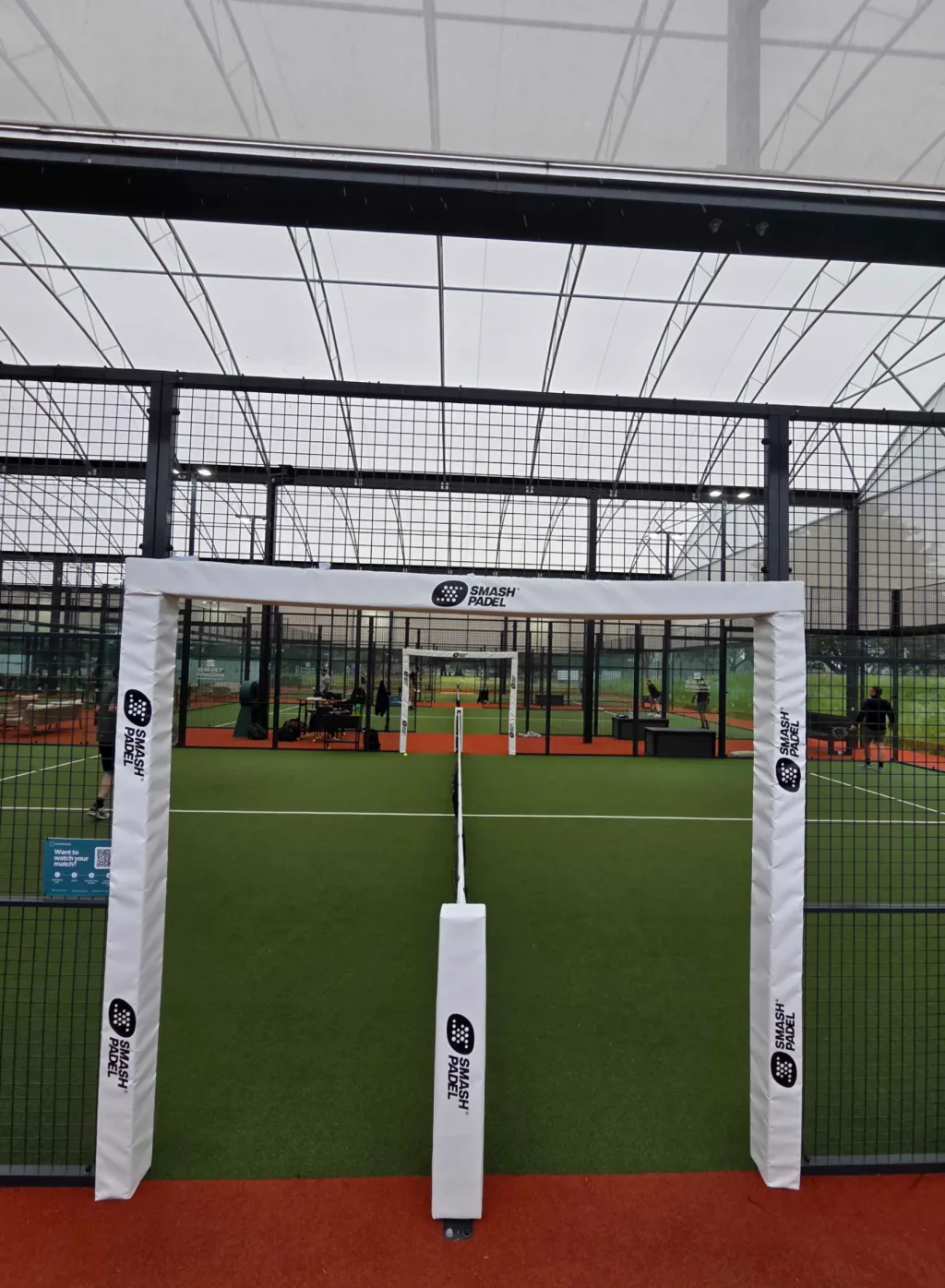 Padel court with white PVC padding reading "SMASH PADEL", green turf, red walkway and wire-mesh walls under a translucent roof.