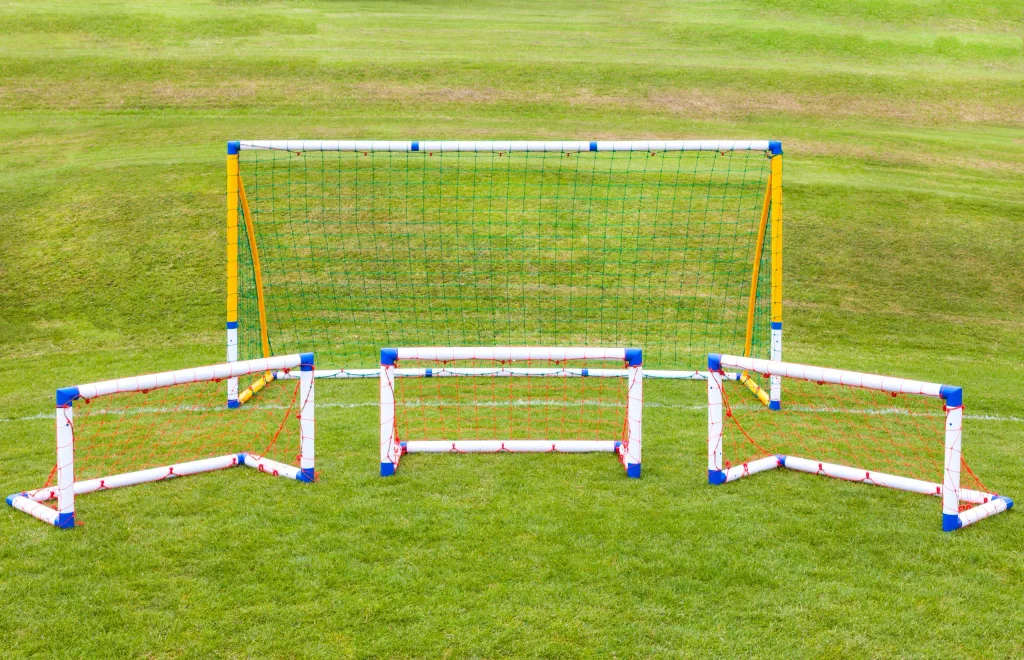 Three small white uPVC training goals with red nets and blue corner caps arranged on grass in front of a larger yellow-framed goal with green net.