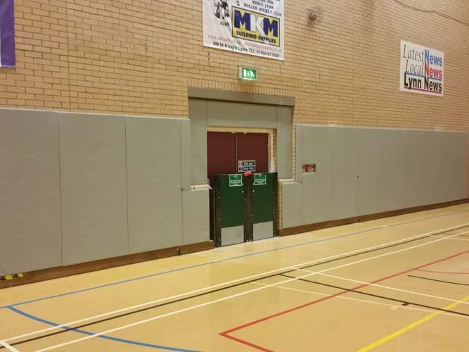 Gym hall with gray padded wall panels and green double fire doors under an exit sign, with a sign reading 'Latest News Local News Lynn News'.