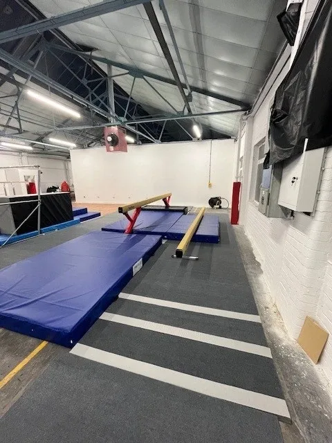 Low balance beam with carbon steel legs positioned over foam landing mats in an indoor gymnastics hall.