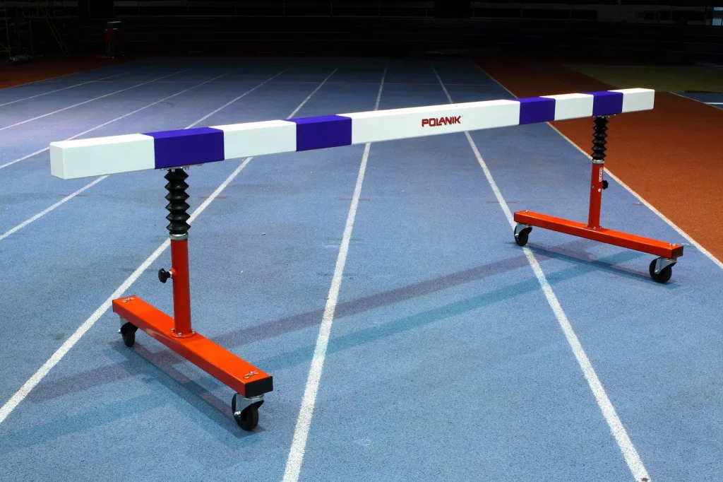 Steeplechase hurdle with a multi-layer wooden beam on orange telescopic base legs with rubber aprons and wheels, positioned on a blue indoor track.