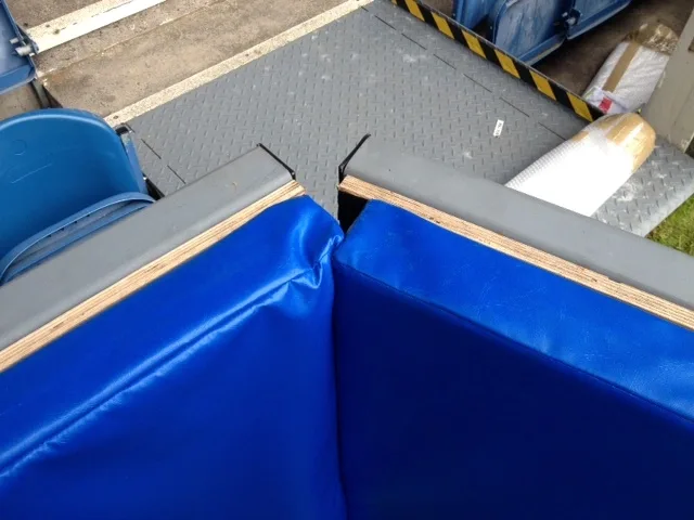 Blue sewn heavy-duty PVC sports pitch safety pads with plywood cores meeting at a corner, grey top edging and stadium seats behind.
