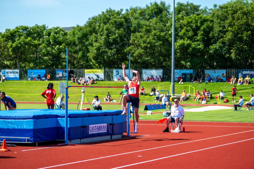 High jumper wearing red bib 88 mid-air over a bar, landing on a blue high-jump mat labeled CANTABRIAN on a red track.