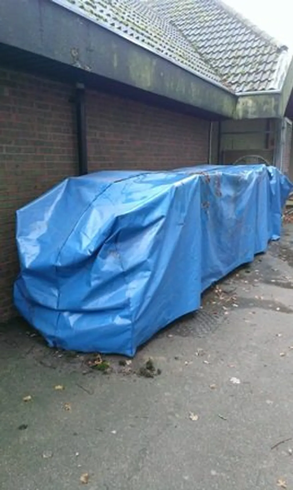 Blue PVC rain cover draped over a long stack of hurdles beside a brick wall under an overhanging tiled roof.
