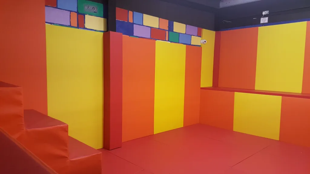 Padded room with red and yellow wall and floor padding, stepped seating on the left, bench seating to the right, and painted brick border reading "ROB'S".