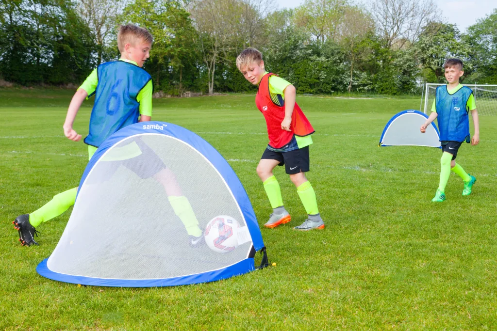 Blue pop-up goal with samba printed on the top in the foreground as three boys in bright training bibs play soccer on a grassy field.