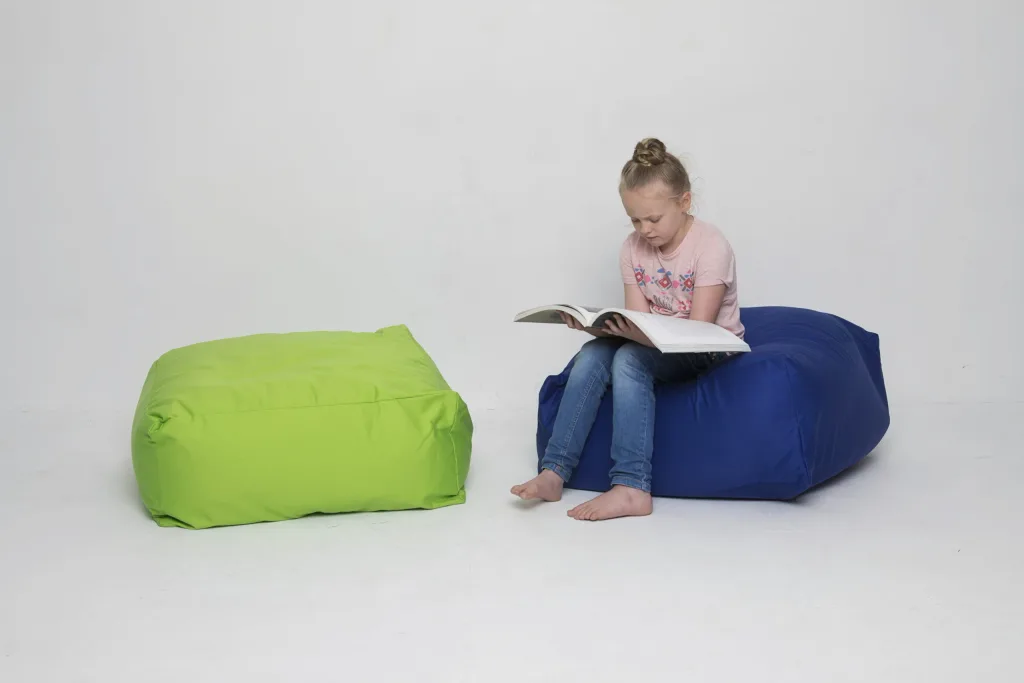 Girl sitting barefoot on a blue cotton child square bean bag reading a book, with a green bean bag beside her.