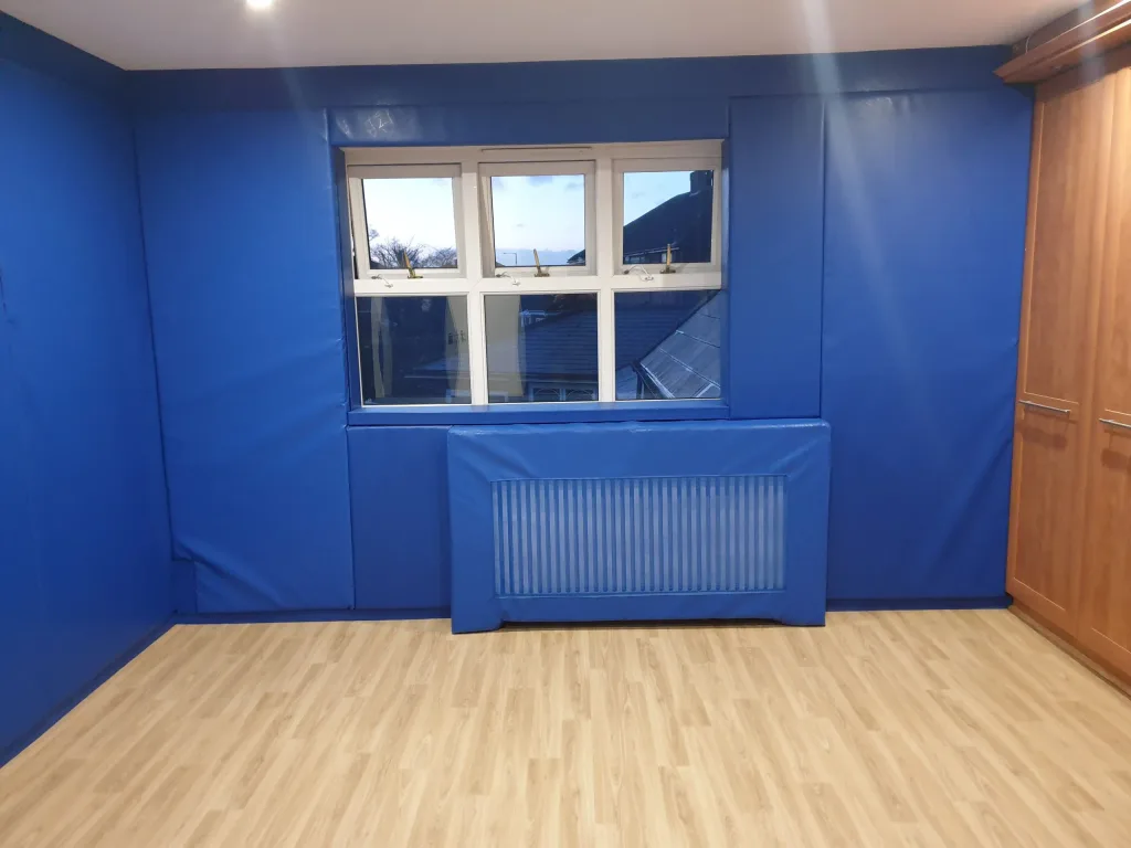 Room with blue padded walls and a padded radiator cover under a central window, light wood laminate floor and a wooden wardrobe at right.