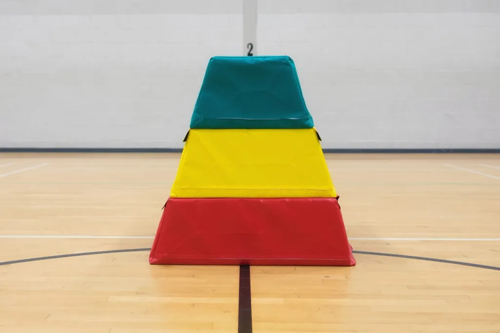 Three-layer foam vaulting box with red base, yellow middle, and teal top, PVC-coated nylon cover, sitting on a gym floor below a wall marked 2.