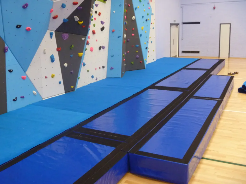 Blue crash mats with heavy-duty carpet tops and thick foam bases beneath angled climbing walls with colorful holds.