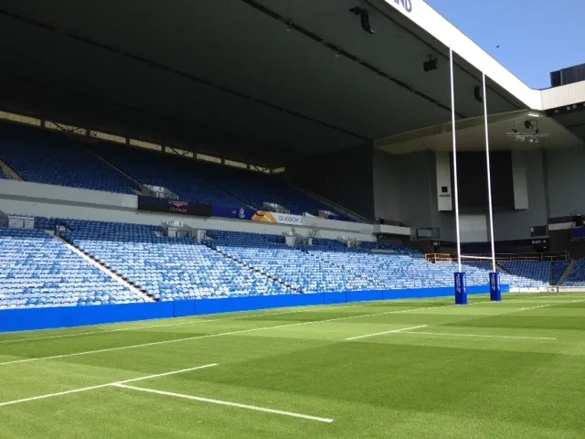 Rugby pitch with green grass, blue safety padding along the sideline and blue padded goalpost protectors in front of empty stands.