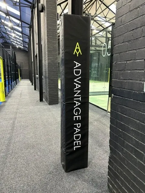 Black padded post beside an indoor padel court with vertical white text ADVANTAGE PADEL and a yellow logo.