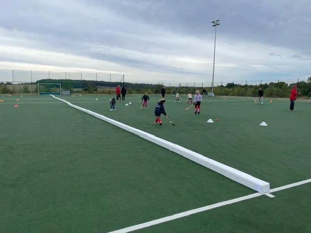 Long white PVC-covered foam hockey pitch divider stretches across green artificial turf while children and coaches practice behind cones.