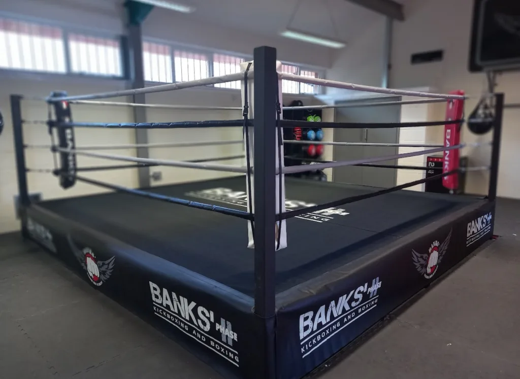 Platform training ring with steel corner posts, black canvas mat, white and black ropes, and padded corner post covers in a gym.