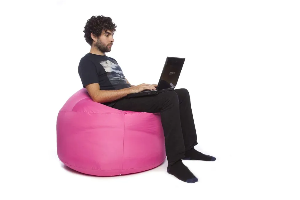 Man seated on a pink polyester bean bag using a laptop against a white background.