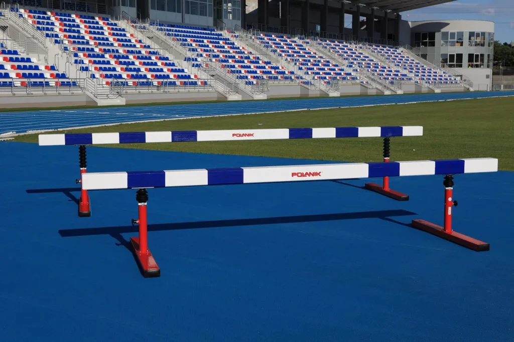 Two white-and-blue wooden steeplechase hurdles on red telescopic legs with rubber aprons on a blue track showing POLANIK on the beams.