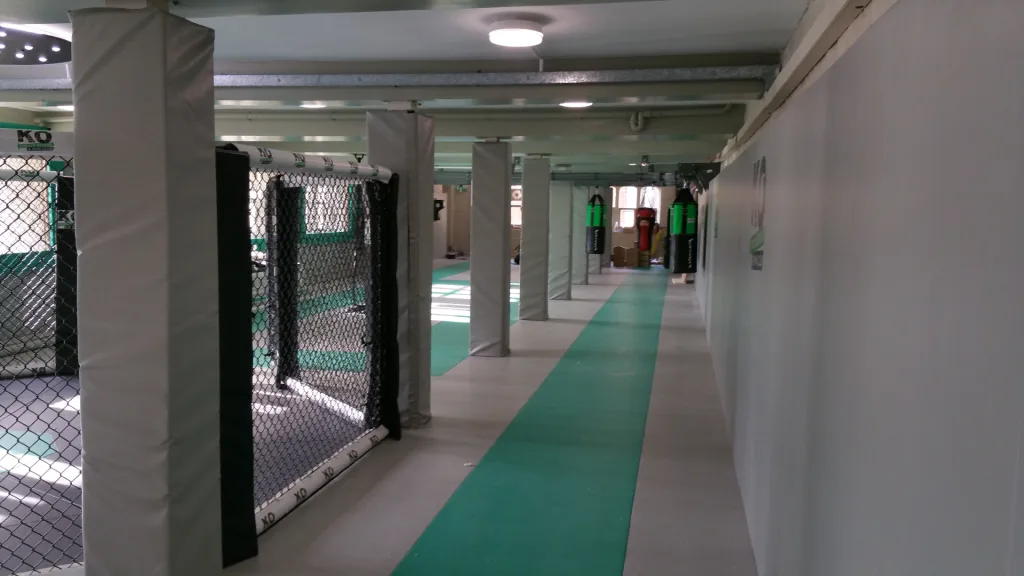 Corridor of an MMA gym with grey padded columns, chain‑link cage, green PVC mats and hanging punching bags, KO on cage pad.