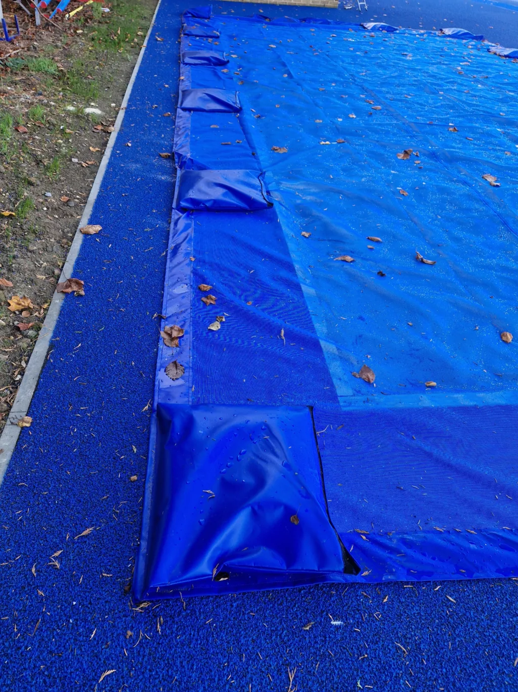 Blue PVC and mesh sandpit cover with edge sleeves and rectangular weight pockets along the left, lying on blue play surfacing with scattered leaves.