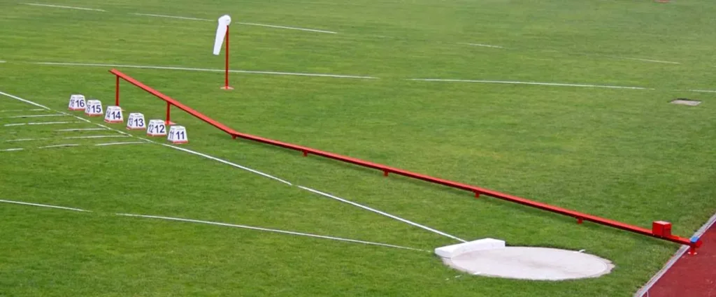 Long powder-painted steel shot put return channel on grass with numbered markers 16 15 14 13 12 11 and a concrete throwing circle.