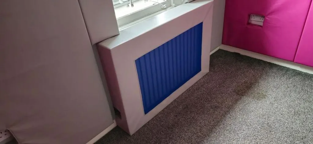 Padded room corner with grey wall panels, a pink padded wall section, a grey padded radiator cover with a blue grille, and grey carpeted floor.