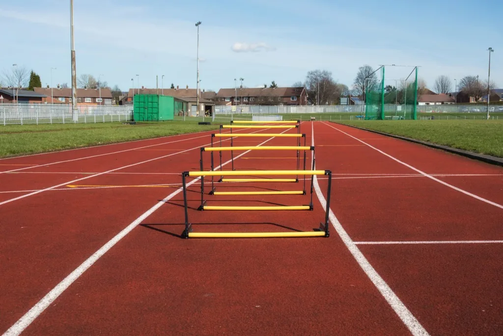 Five yellow training hurdles with black frames lined up down the center of a red outdoor running track.