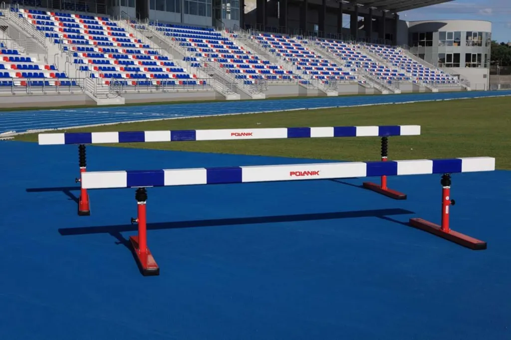 Two adjustable steeplechase hurdles with white-and-blue striped multi-layer wooden beams on red telescopic legs standing on a blue track before empty stadium stands.
