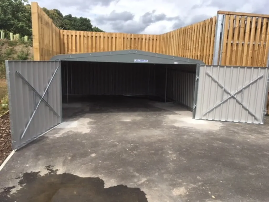 Grey galvanised steel hurdle garage with open double doors on a paved area, backed by a tall wooden fence.