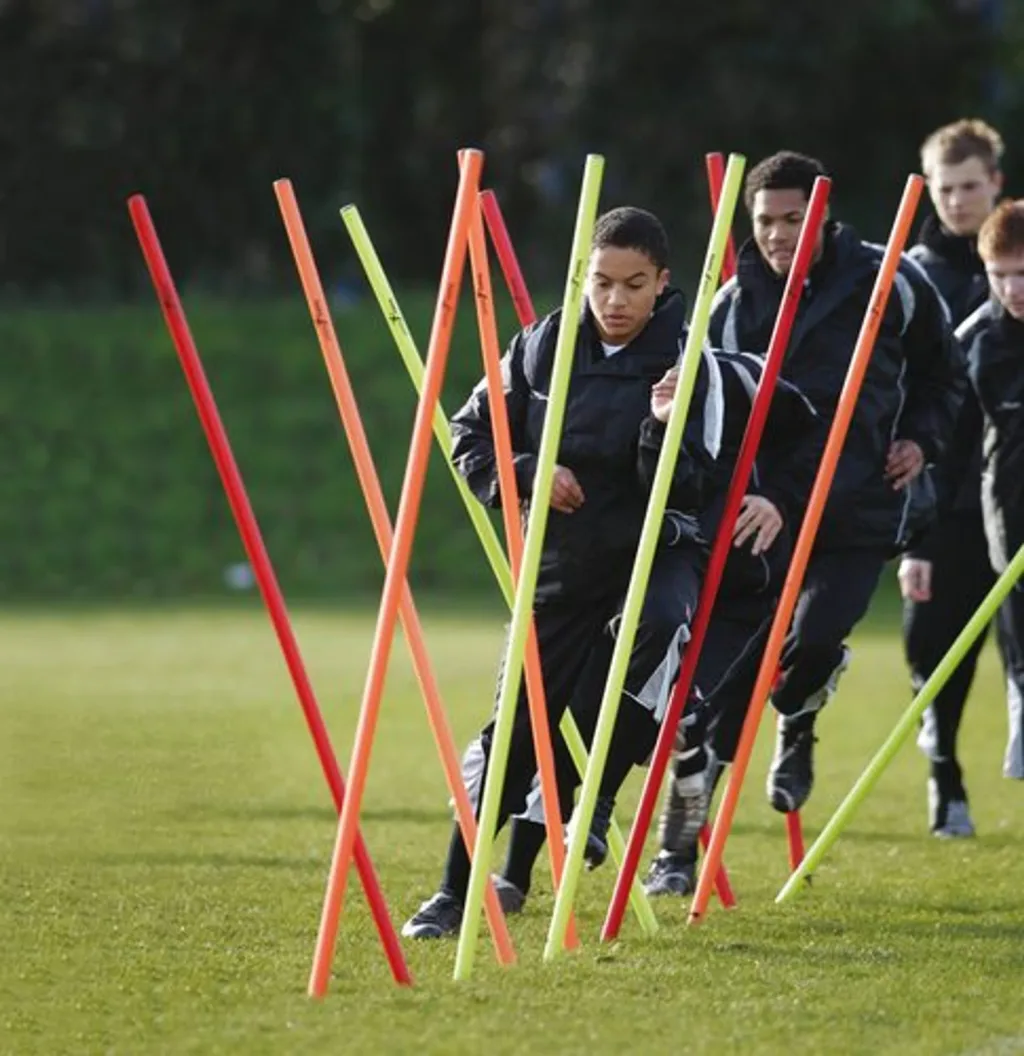 Group of players in black tracksuits running through fluo yellow, fluo orange, and red boundary poles set in grass.