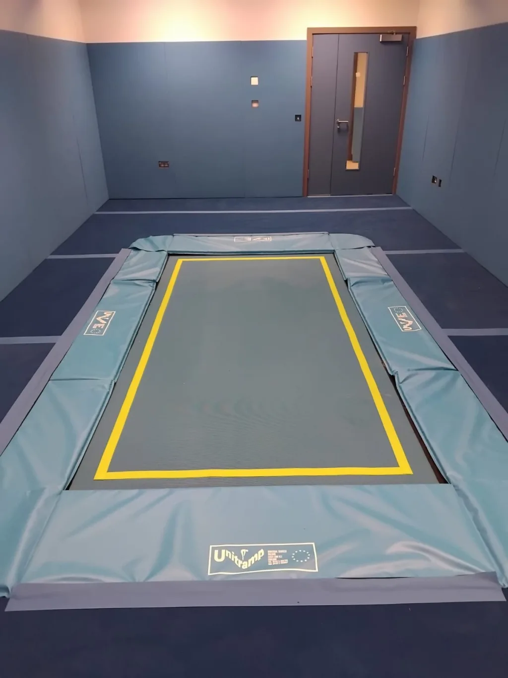 Recessed rectangular trampoline with a yellow border and light blue surround padding labeled "Unitramp" in a blue padded therapy room.