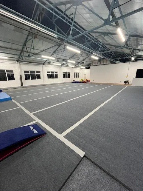 Light grey carpet roll-out gymnastics mats with white lane lines in a large gym hall, blue mat in foreground labeled 'REBEL'.