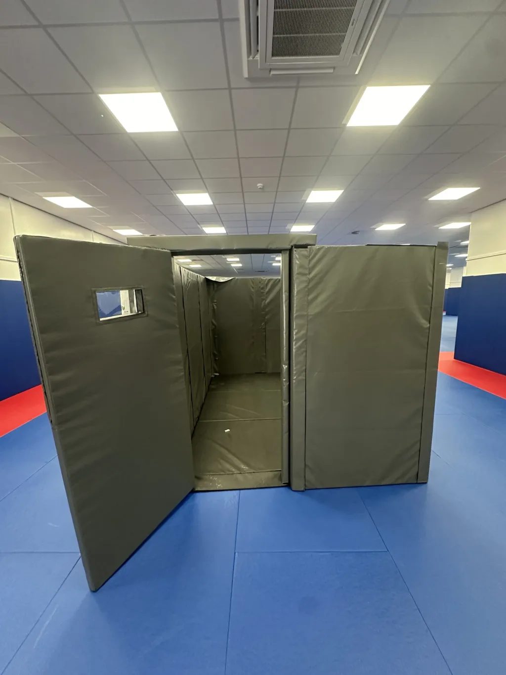 Modular foam custody cell covered in heavy-duty PVC with an open door and small viewing window, set on blue training mats in a hall.
