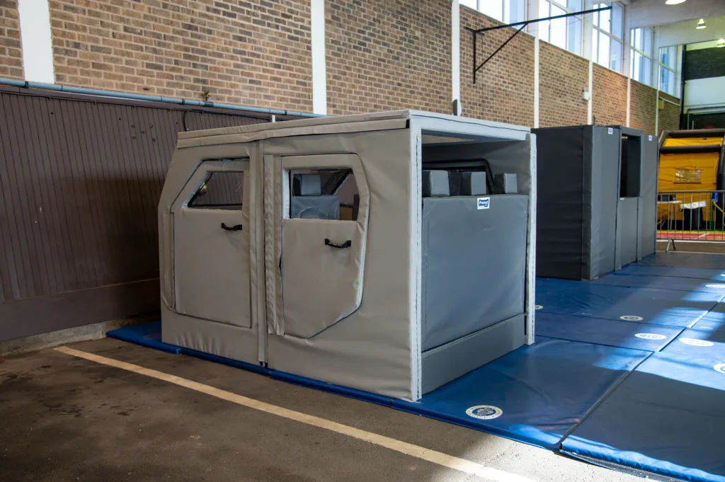 Grey PVC-covered foam training car with window openings and door flaps, label reading Promat, sitting on blue mats inside a gym-like hall.
