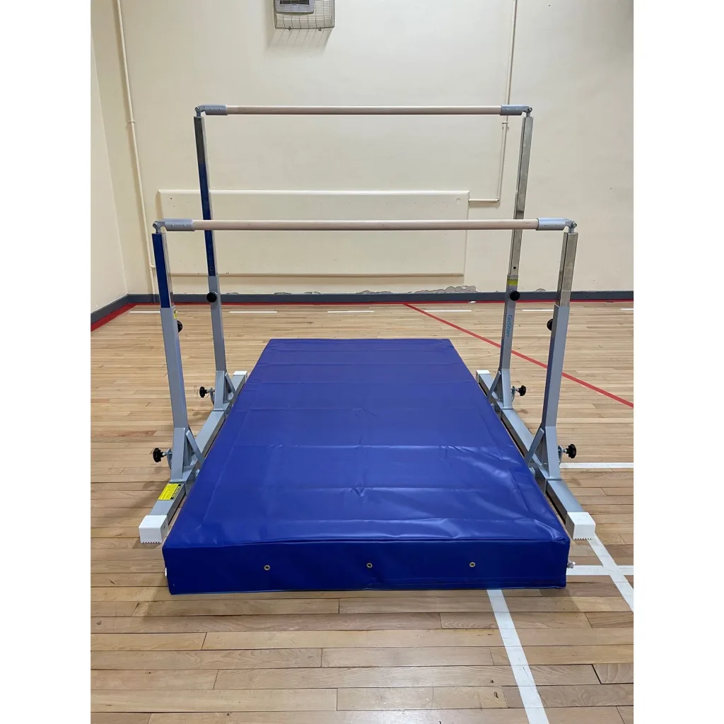 Adjustable children's uneven bars with wooden rails on a carbon steel frame set over a blue crash mat on a gym wooden floor.
