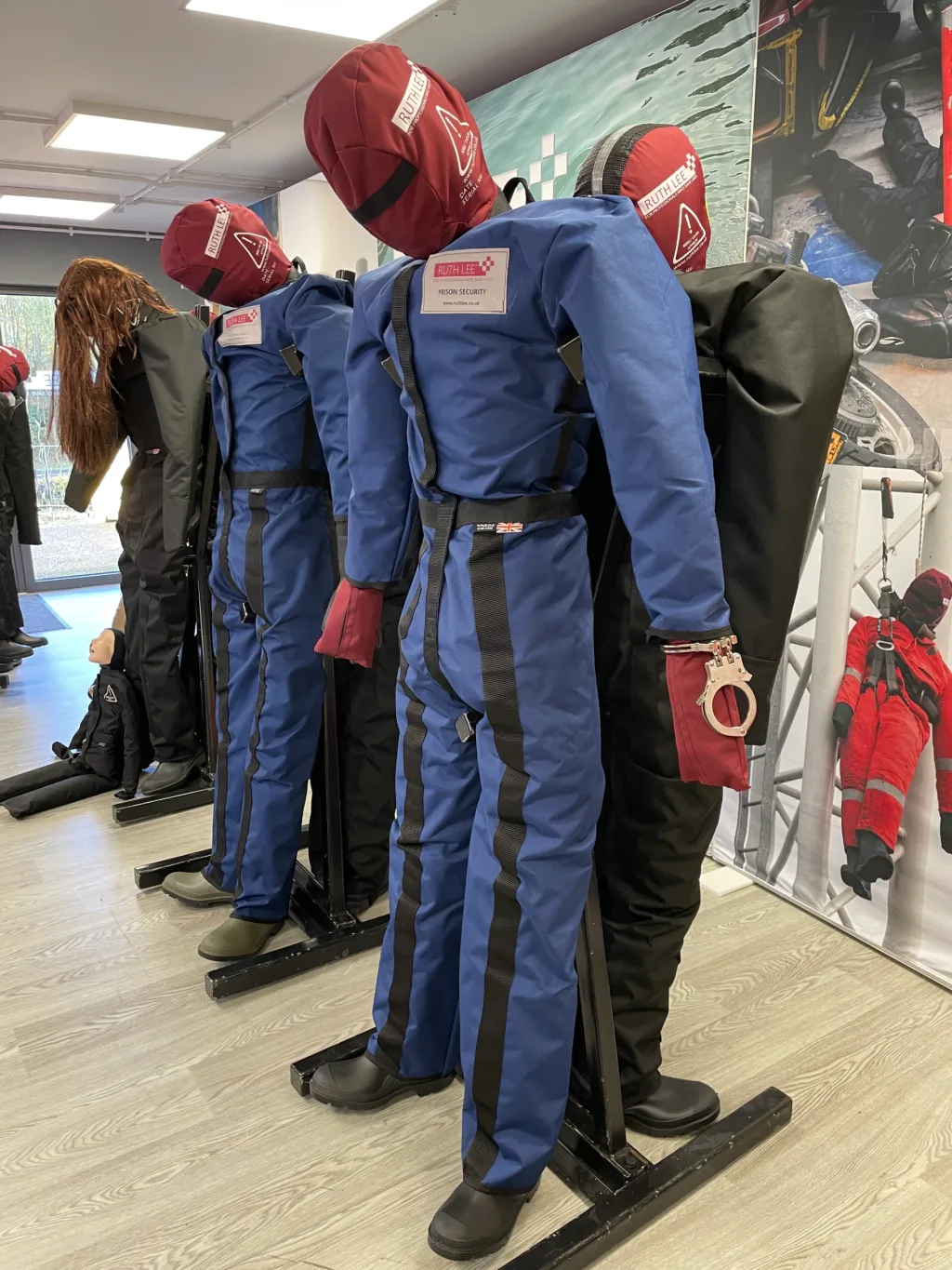 Three prisoner security manikins wearing blue flame-retardant polyester overalls and red hoods, one with handcuffs on a wrist, displayed on black stands in a showroom.
