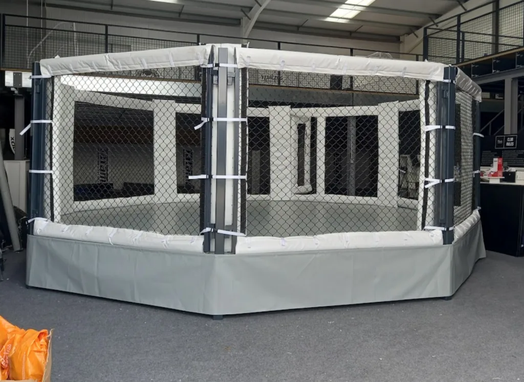 Octagonal competition cage with chain-link sides, white padded top, grey side skirt and powder-coated steel frame on a raised deck inside a showroom.