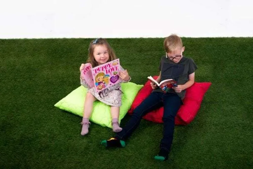 Two children reading books while sitting on lime and red padded floor cushions on green grass, girl holding a magazine titled "PUPPY LOVE".