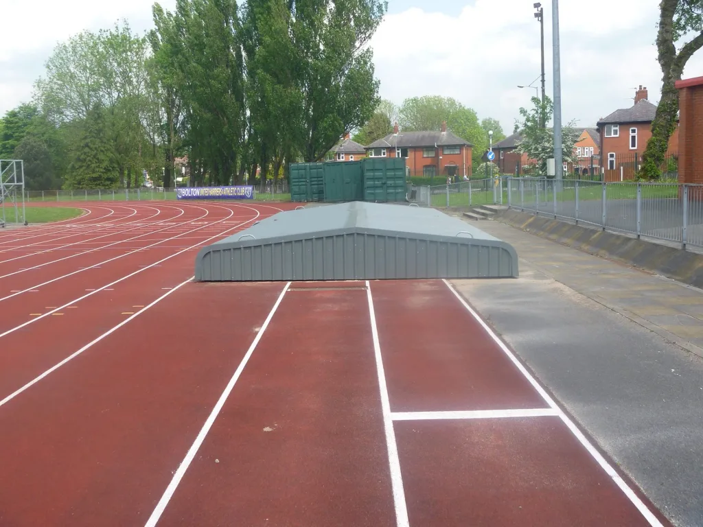 Steel wheelaway sandpit cover over a long jump pit at the end of a red running track, two gray steel sections with handles beside a paved walkway.