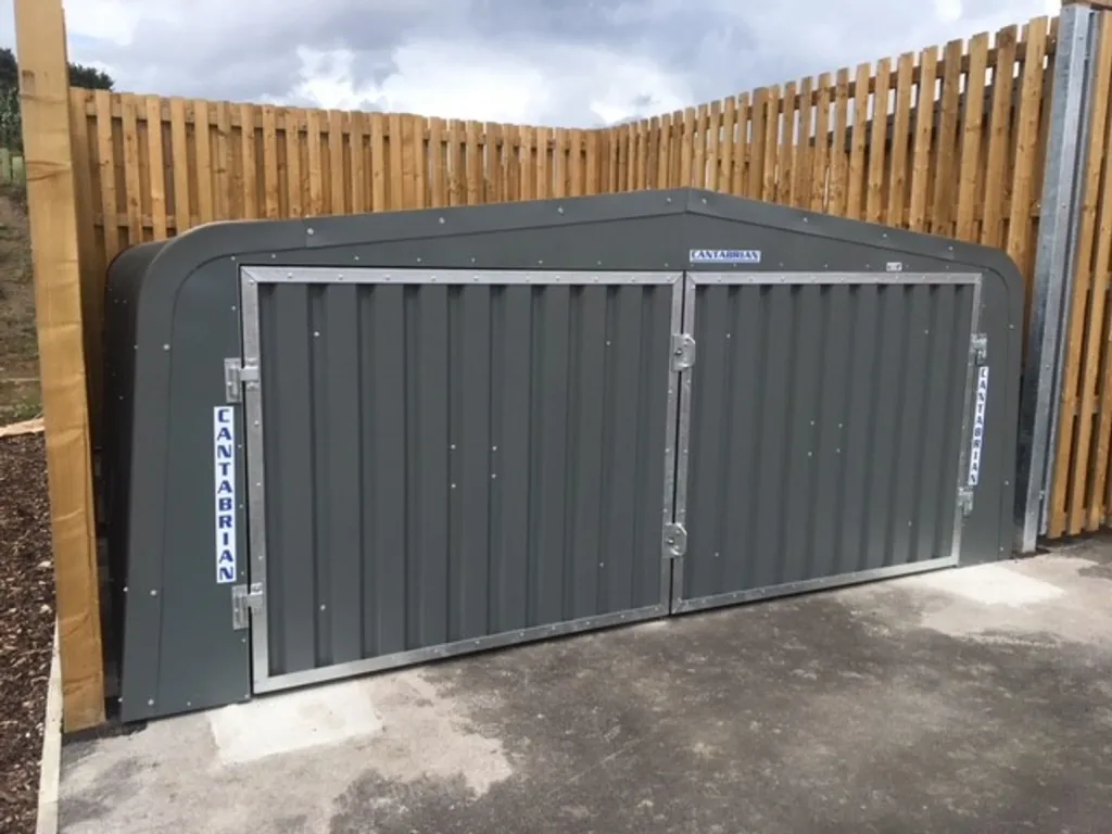 Galvanised steel hurdle garage with double corrugated doors and steel frame set on concrete against a wooden fence.
