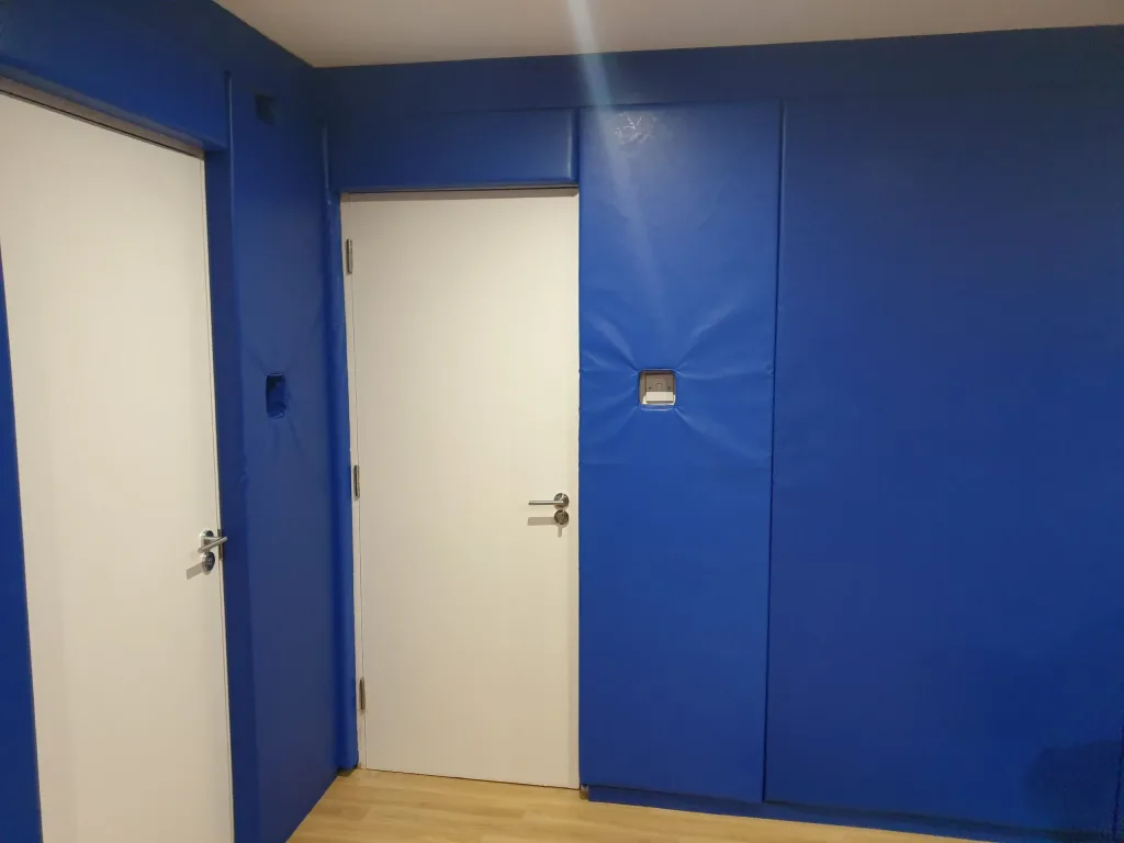 Blue padded walls lining a room with two white doors and a wood floor, one pad has a small square access cutout.