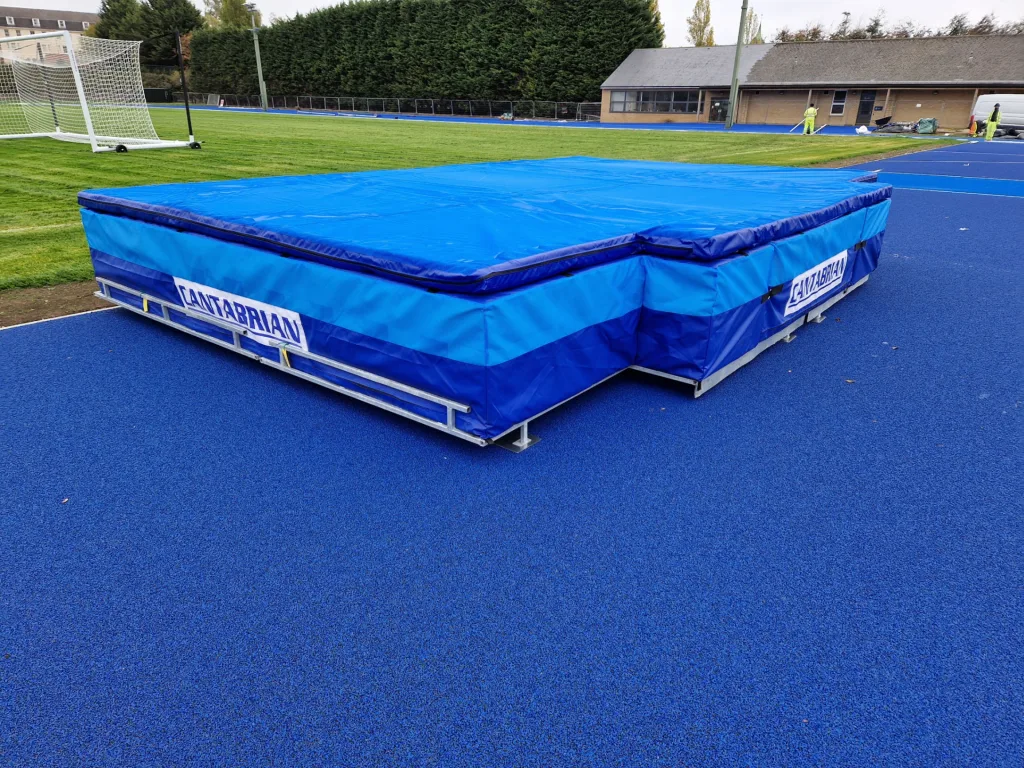 Galvanised steel platform supporting blue high-jump landing mats with guardrail, placed on a blue athletics track.