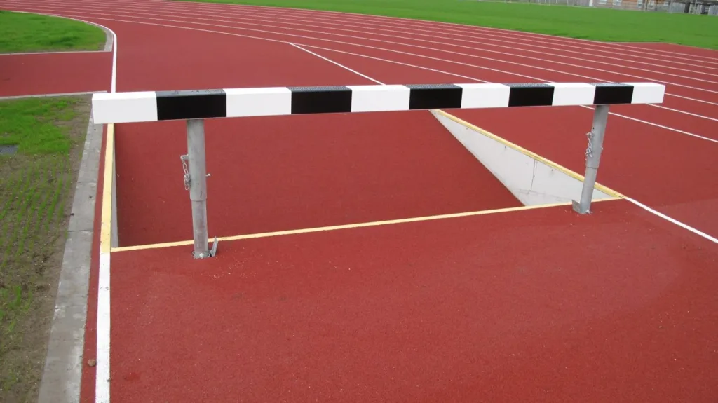 White-and-black striped wooden water jump barrier on two galvanised metal uprights spanning a red track water pit.