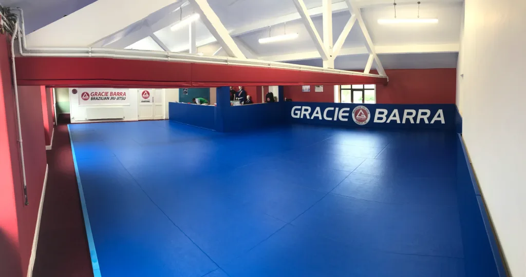 Royal blue MMA mats cover a training hall with white roof beams and a wall reading "GRACIE BARRA".