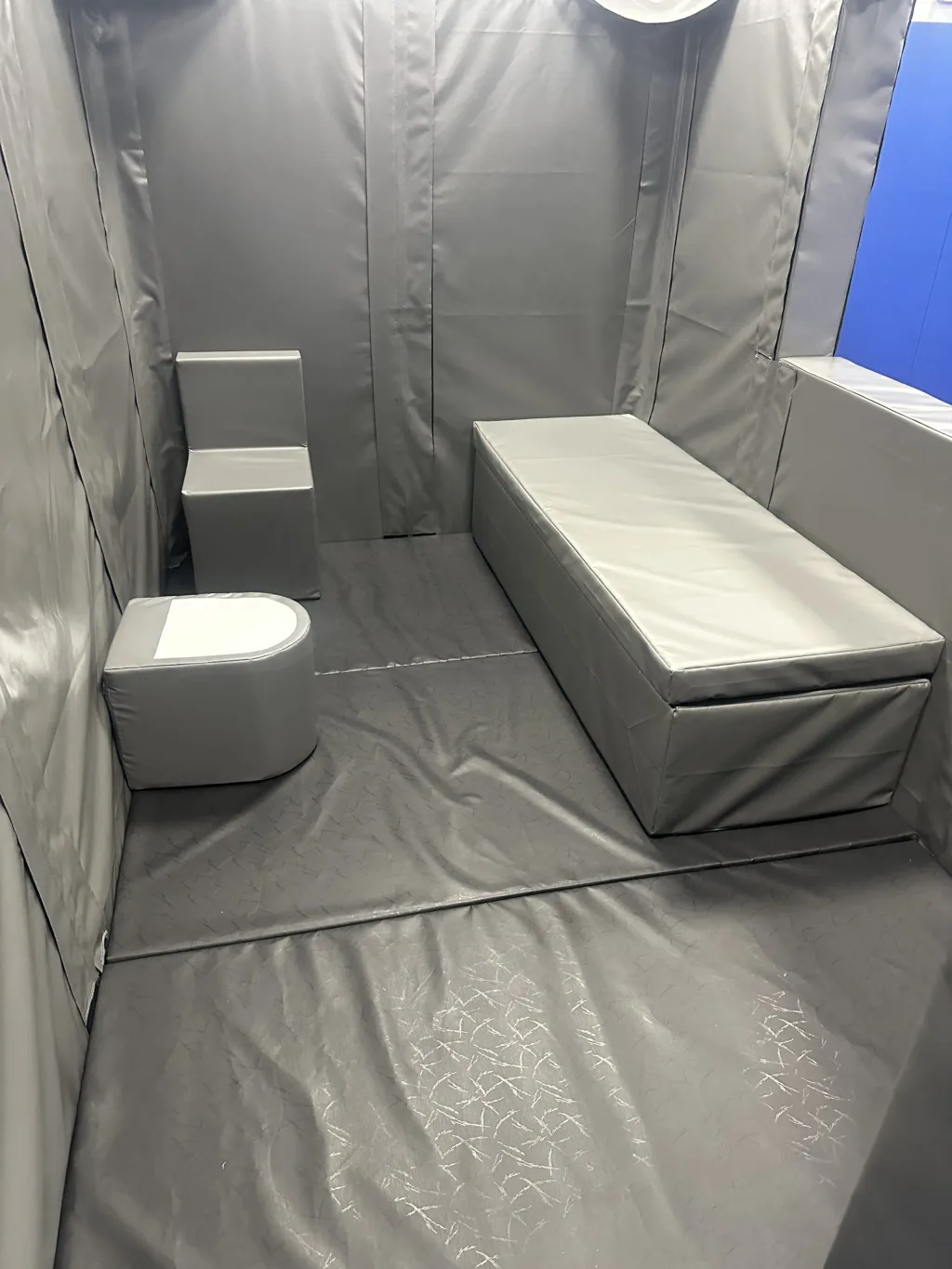 Padded custody cell interior with grey PVC walls, a long padded bench/bed, a foam toilet and a small block seat on a grey padded floor.