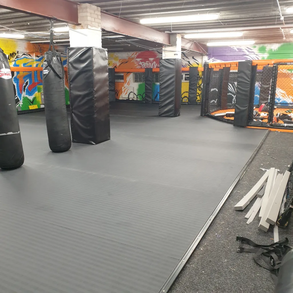 Large black PVC roll-out tatami-style mat covering a martial arts gym floor with hanging heavy bags, padded pillars and an orange cage at the back.