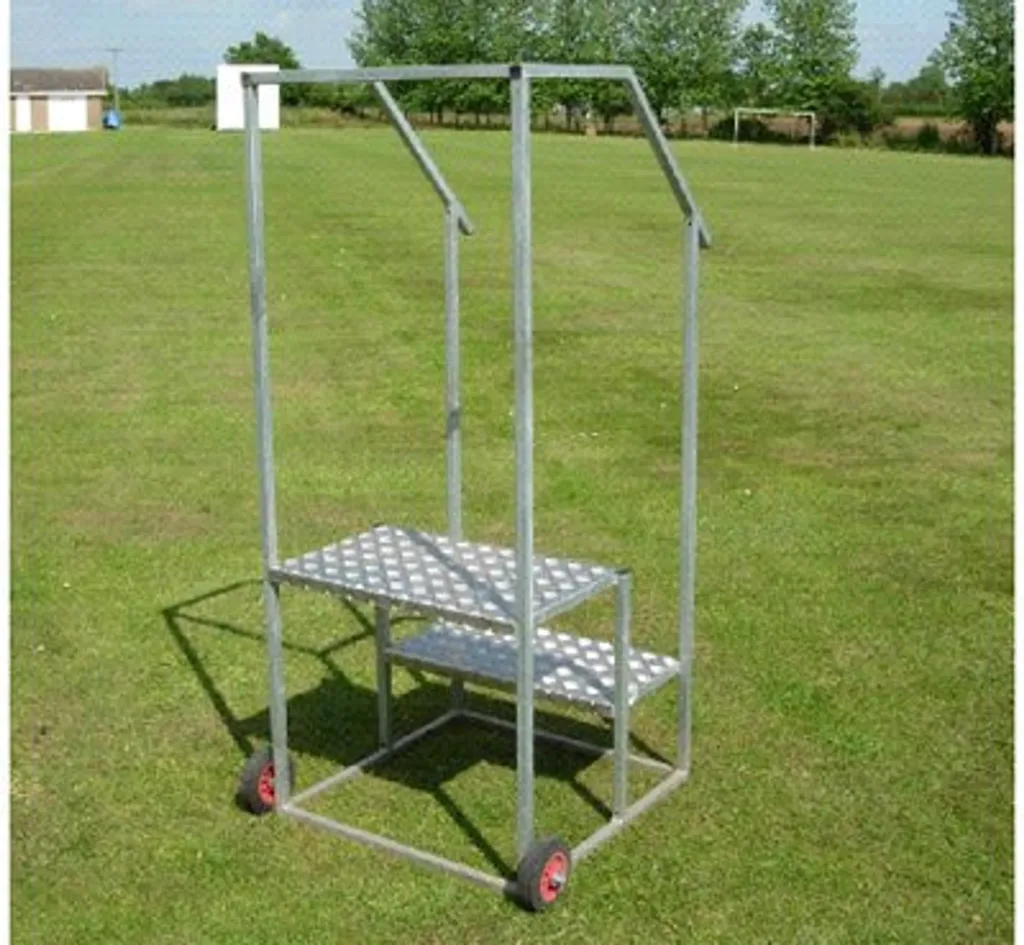 Galvanized steel and aluminium two-step starters rostrum with five-bar treadplates, handrails and two wheels on a grassy sports field.