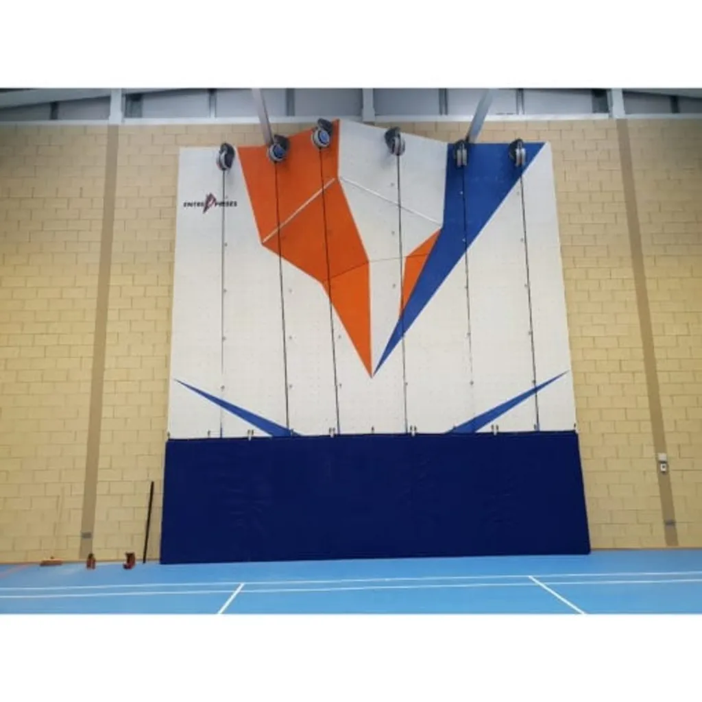 Blue folded hanging mats mounted along the base of a white indoor climbing wall with orange and blue geometric panels and hanging ropes above a pale blue sports floor.